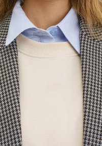 Brązowa i niebieska marynarka w kratę na beżowym swetrze, z jasnoniebieską koszulą z kołnierzykiem pod spodem. Teksturowane tkaniny i warstwowy design.