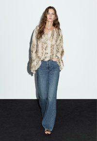 Löst sittande beige blus med ormtryck, har volangärmar. Kombineras med hög midja blå utsvängda jeans och sandaler.