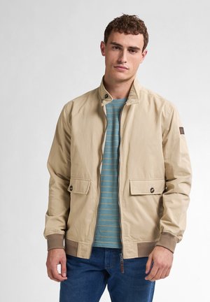 Giacca leggera beige con colletto alto, zip frontale, due tasche a toppa e polsini a coste. Indossata sopra una camicia a righe color teal e jeans blu.