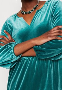 Tyrkysové sametové šaty s výstřihem do V, nařasenými dlouhými rukávy a splývavým siluetam. Doplněné stříbrným náhrdelníkem se smaragdovými detaily.