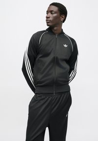 Juodas sportinis švarkelis su baltomis juostelėmis rankovėse, užtrauktuku priekyje ir mažu logotipu ant krūtinės. Derinamas su derančiomis juodomis sportinėmis kelnėmis.