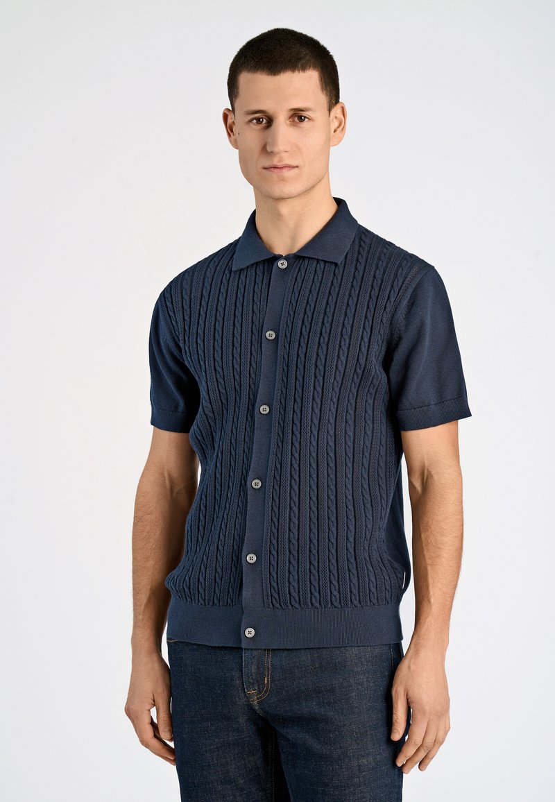 Tumedat lühikeste varrukatega kudumispullover, millel on nööpide sulgemine ja soonikkoes varrukate ääred. Kombineeritud tumedate teksadega.