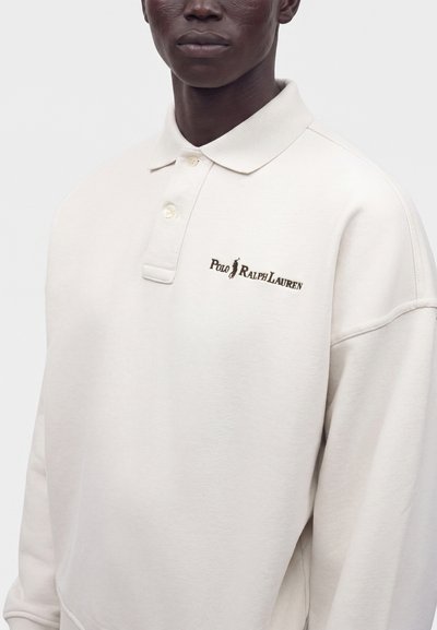 Άνδρας φοράει λευκό μακρυμάνικο πόλο Ralph Lauren με γιακά και δύο κουμπιά, με κέντημα λογότυπου στο στήθος.
