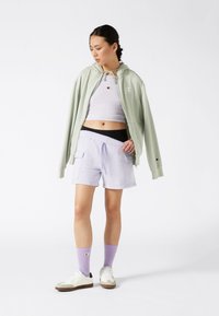 Giacca con cappuccio verde chiaro con zip, top corto lavanda, pantaloni corti morbidi lavanda con tasche e scarpe da ginnastica bianche con suole nere e calzini viola.