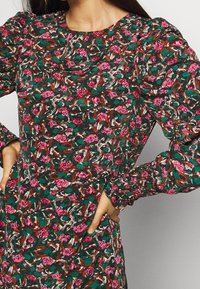 Blumenkleid in Dunkelgrün mit rosa und weißen Mustern. Verfügt über Puffärmel und geraffte Manschetten. Aus leichtem Stoff gefertigt.
