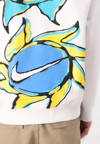 Bela puhovka s posadko v belem bombažu, ki vključuje drzne, pisane sončne in krožne vzorce v modri, rumeni in črni barvi z akcentom Nike logotipa.