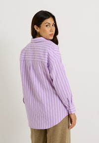 Camisa a rayas morado claro con líneas verticales blancas, con cuello abotonado, mangas largas y un dobladillo ligeramente curvado en la parte posterior.