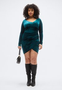 Φόρεμα από βελούδο σε τιρκουάζ με μακριά μανίκια, με σχεδιασμό τύπου κορσέ και ψηλή σκίσιμο. Συνδυάζεται με μαύρες μπότες μέχρι το γόνατο και μικρή μαύρη τσάντα χειρός.