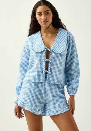 Kvinde med langt krøllet hår iført blå og hvide ternet bluse med lange ærmer, stor krave og sløjfer, kombineret med matchende shorts med flæser.