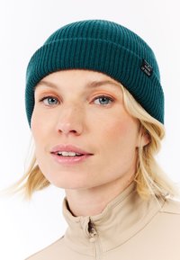 Tyrkysová pletená čepice s žebrovaným vzorem, s ohrnutým lemem a malou černou etiketou. Nositelná s krémovým zipovým svrškem. Hladká textura a těsný střih.