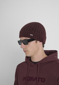 Burgundska rebrasta pletena kapa, uparena s crnim pravokutnim naočalama. Model nosi burgundsku duksericu s natpisom "MORATO" u odgovarajućoj boji.