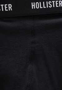 Μαύρο ύφασμα με ορατή κάθετη ραφή και μερική θέα στη μαύρη μέση που φέρει λευκή επιγραφή "Hollister".