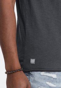 T-shirt corto grigio scuro con finitura testurizzata, dotato di un'etichetta grigia del marchio sull'orlo. Indossato con jeans strappati.