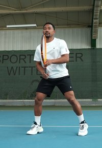 Athletischer Mann in weißem, feuchtigkeitsableitendem Shirt und schwarzen Shorts, der einen orangefarbenen Tennisschläger auf einem blauen Platz mit grünem Hintergrund hält.