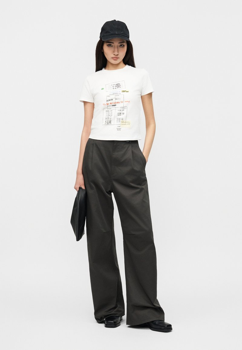 Jeune femme portant une casquette noire, un t-shirt court blanc à motif graphique, un pantalon noir ample, des chaussures noires, tenant une pochette noire, debout devant un fond gris.