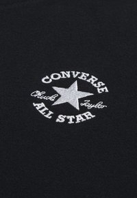 Svart tyg med en cirkulär vit broderad logotyp med en stjärna och texten "CONVERSE Chuck Taylor All Star" i fet stil.