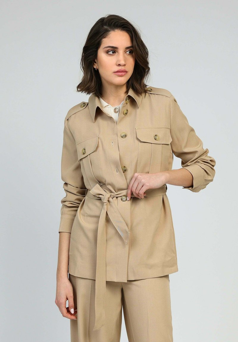 Femme ajustant la ceinture d'une veste beige à boutons avec poches poitrine, assortie à un pantalon, devant un fond gris clair uni.