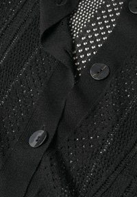 Schwarze Strickjacke mit einem strukturierten Muster aus offenen Maschen und Stoffknöpfen. Die Zusammensetzung umfasst weiches Material und dezente Details.