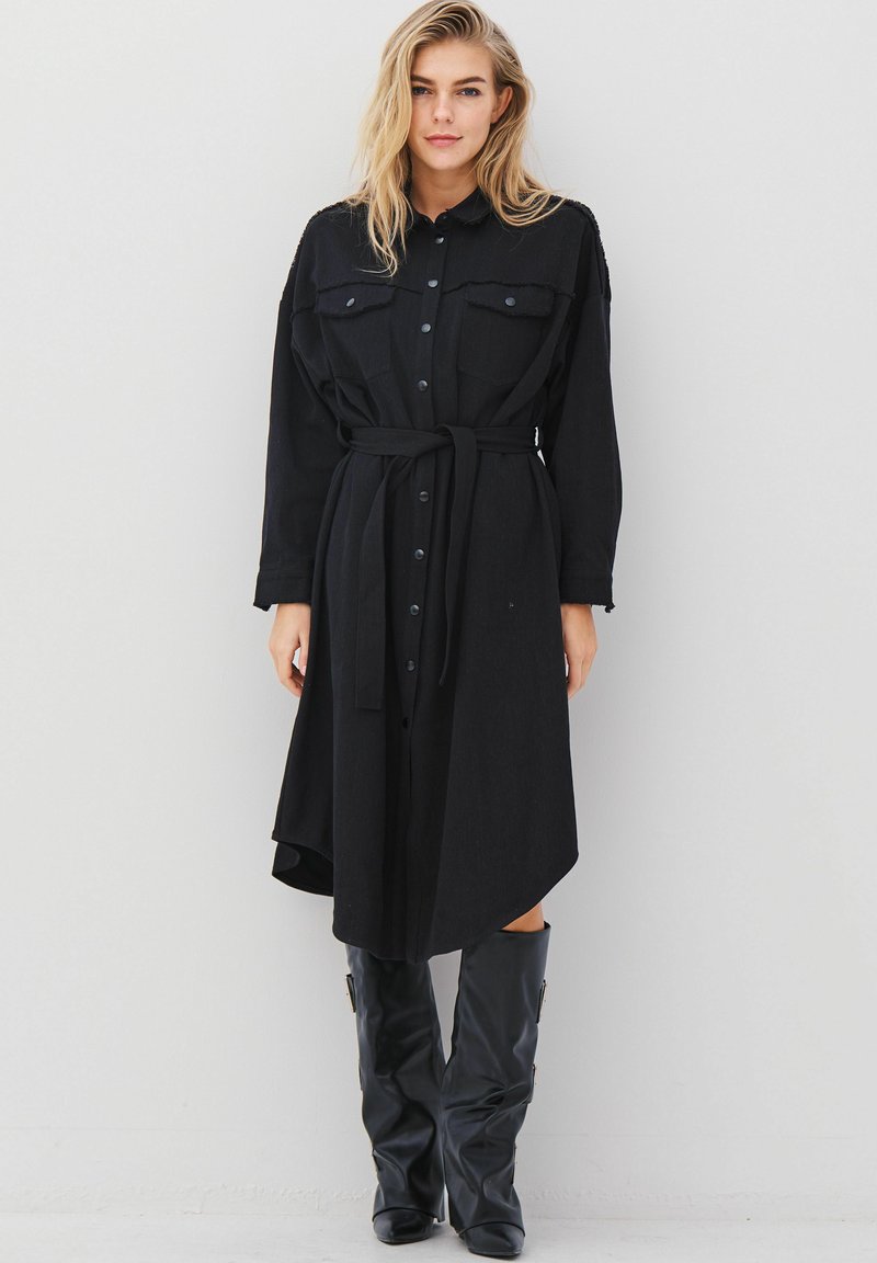 Femme aux cheveux blonds portant une robe noire boutonnée avec ceinture et des bottes en cuir noires montantes jusqu'aux genoux, debout contre un mur clair uni.