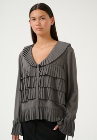 Blusa grigia plissè con scollatura a V profonda e ruffles sovrapposti. Presenta chiusura con bottoni e dettagli con cravatta sulle maniche. Tessuto morbido e strutturato.
