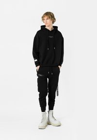 Sudadera negra con capucha y logo frontal, combinada con pantalones jogger negros con cremalleras en los bolsillos. Lleva puestas botas de combate blancas.