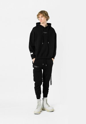 Sudadera negra con capucha y logo frontal, combinada con pantalones jogger negros con cremalleras en los bolsillos. Lleva puestas botas de combate blancas.