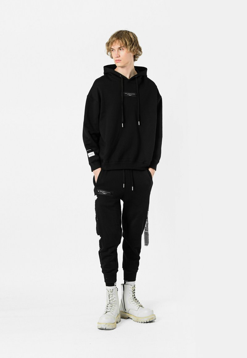 Sudadera negra con capucha y logo frontal, combinada con pantalones jogger negros con cremalleras en los bolsillos. Lleva puestas botas de combate blancas.