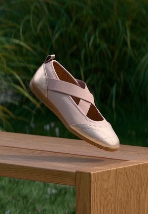 Ballerine rose pâle avec des brides élastiques croisées flottant au-dessus d'un banc en bois en plein air avec des plantes vertes en arrière-plan.