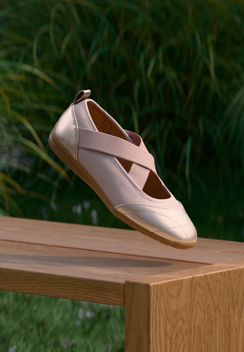 Παλλό ροζ μπαλέτο μπαλαρίνα με ελαστικές λουρίδες που διασταυρώνονται, αιωρούμενο πάνω από ένα ξύλινο παγκάκι σε εξωτερικό χώρο με πράσινα φυτά στο παρασκήνιο.