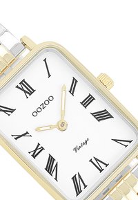 Montre rectangulaire en or et en argent avec un cadran blanc, des chiffres romains noirs et des aiguilles en or. Présente un bracelet en métal texturé.