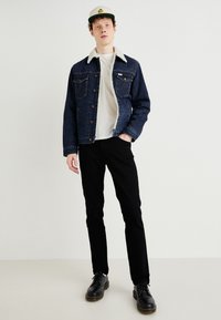 Jachetă din denim cu guler din fleece, de culoare albastru închis, asortată cu un tricou alb și pantaloni negri. Pantofi negri cu cusături galbene, stând pe o podea deschisă.