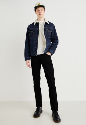 Jeans jakna s flis ovratnikom u tamnoplavoj boji, u kombinaciji s bijelom majicom i crnim hlačama. Crne cipele s žutim šavovima, stojeći na svijetloj podlozi.