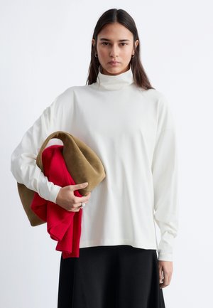 Ung kvinde med langt mørkt hår iført en hvid rullekrave og sort nederdel, der holder rødt og brunt foldet tøj i venstre arm.