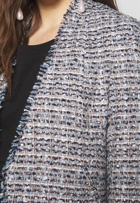 Blazer in tweed caratterizzato da una miscela di fili blu, navy e beige, con una finitura testurizzata e bordi sfrangiati, abbinato a un top nero.