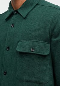 Zielona koszula-kurtka z mieszanki wełny o teksturowanym wykończeniu, z guzikami, kołnierzem i kieszonką na klapie po lewej stronie.