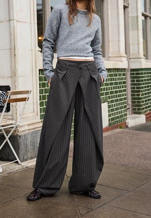 Vrouw draagt een grijze cropped trui en wijd uitlopende donkergrijze pantalon met krijtstrepen, staand buiten bij een betegelde muur en een klapstoel.