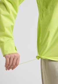 Limegrüner, leichter Jacke mit Reißverschluss und verstellbarem Saum. Sie verfügt über eine Hand, die den Kordelzug am unteren Ende zieht, um eine enge Passform zu gewährleisten.