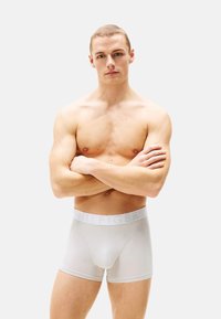 Boxers blancos hechos de tela elástica con una cinturilla suave que presenta el logo "HILFIGER" en gris claro. Modelo posando con los brazos cruzados.