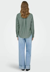 Camisa verde de manga larga con un patrón texturizado, dobladillo curvado y puños abotonados, combinada con vaqueros anchos de pierna en azul claro.