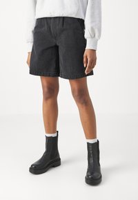 Černé džínové kraťasy s rovnostranným střihem a volným volným střihem, se středně vysokým pasem, doplněné černými kotníkovými botami a bílými ponožkami.