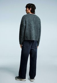 Grauer, flauschiger Pullover mit kurzem Schnitt, kombiniert mit lockeren, dunkelblauen Jeans. Gestylt mit hellen Schuhen, von hinten betrachtet.