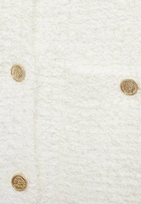 Weißer, strukturierter Stoff mit einer samtigen Oberfläche, der an der Vorderseite vier goldene Knöpfe aufweist. Die Knöpfe sind mit einem dekorativen Muster geprägt.