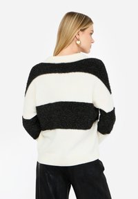 Maglione a righe bianche e nere con texture soffice, caratterizzato da una vestibilità ampia, scollo largo e tessuto tricot morbido. Orecchini a forma di cuore dorati.
