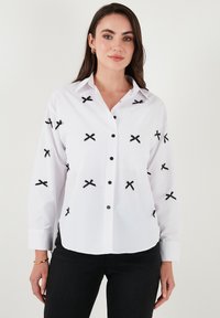 White button-up shirt with black cross embroidery, long sleeves, buttoned cuffs, and black buttons. Smooth texture, classic collar design.