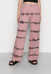 Rosa vida byxor med svarta tie-dye-ränder, tillverkade av bomull, fem fickor, bälteshällor och en knappstängning framtill. Bärs med svarta sandaler.