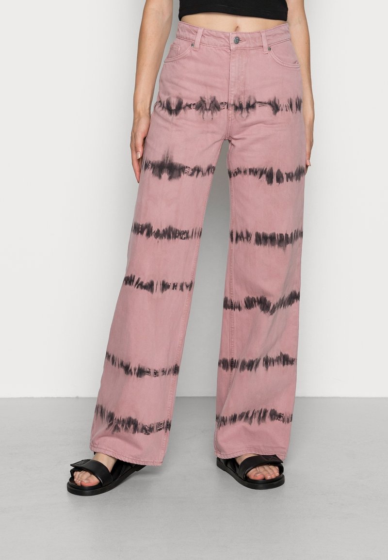 Rosa vida byxor med svarta tie-dye-ränder, tillverkade av bomull, fem fickor, bälteshällor och en knappstängning framtill. Bärs med svarta sandaler.