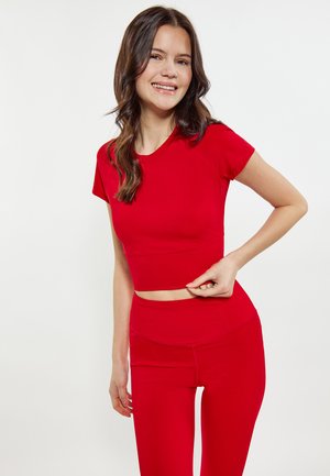 Nuori nainen hymyilee, pukeutuneena punaiseen istuvaan crop-toppiin ja korkeavyötäröisiin legginsseihin, pitää toisen käden topin helmassa.
