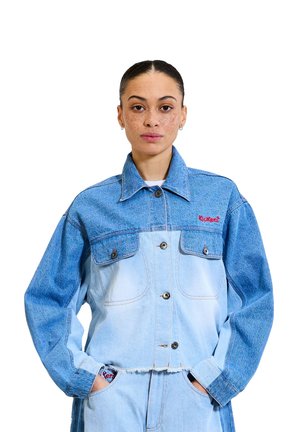 Giubbotto in denim azzurro chiaro con tasche sul petto di un blu più scuro, grandi bottoni e orlo sfilacciato. Logo ricamato rosso sul lato sinistro del petto.