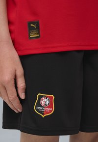 Punane jalgpallipaita tekstureeritud kangast, musta sildiga. Mustad lühikesed püksid, millel on Stade Rennaise klubi logo, kullast ja punasest detailid, sile tekstuur.