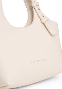 Kremowa skórzana torebka o zaokrąglonym kształcie, z regulowanym paskiem i złotym tłoczonym logo "TOMMY HILFIGER" u podstawy.